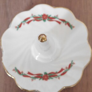 May include: White ceramic ring holder with a scalloped edge and gold trim. The ring holder features a central post and is decorated with red ribbon and green holly accents. A decorative piece for a dresser or vanity.
