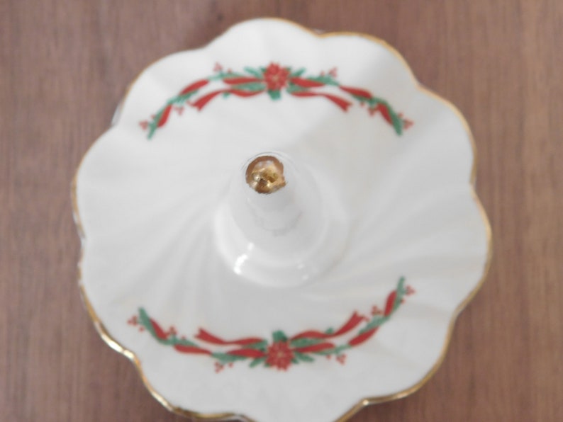 May include: White ceramic ring holder with a scalloped edge and gold trim. The ring holder features a central post and is decorated with red ribbon and green holly accents. A decorative piece for a dresser or vanity.