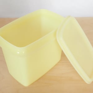 May include: A yellow plastic container with a lid. The container is rectangular and has a smooth surface. The lid is slightly curved and has a smooth surface.