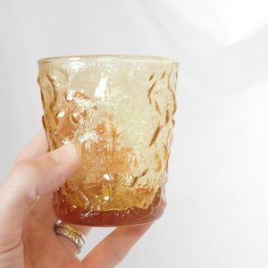 May include: A clear amber glass tumbler with a textured, embossed design. The glass has a rounded bottom and a straight, slightly flared top.