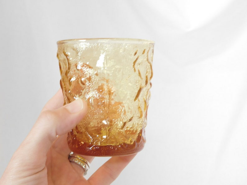 May include: A clear amber glass tumbler with a textured, embossed design. The glass has a rounded bottom and a straight, slightly flared top.