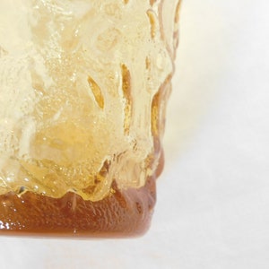 May include: A close-up of a clear glass vase with a textured surface. The vase is amber in color and has a rounded bottom.
