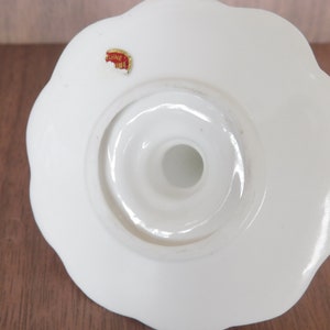May include: A white ceramic candle holder with a scalloped edge. The base has a circular opening and a small sticker that reads "Genuine China". The candle holder is designed to hold a single candle.