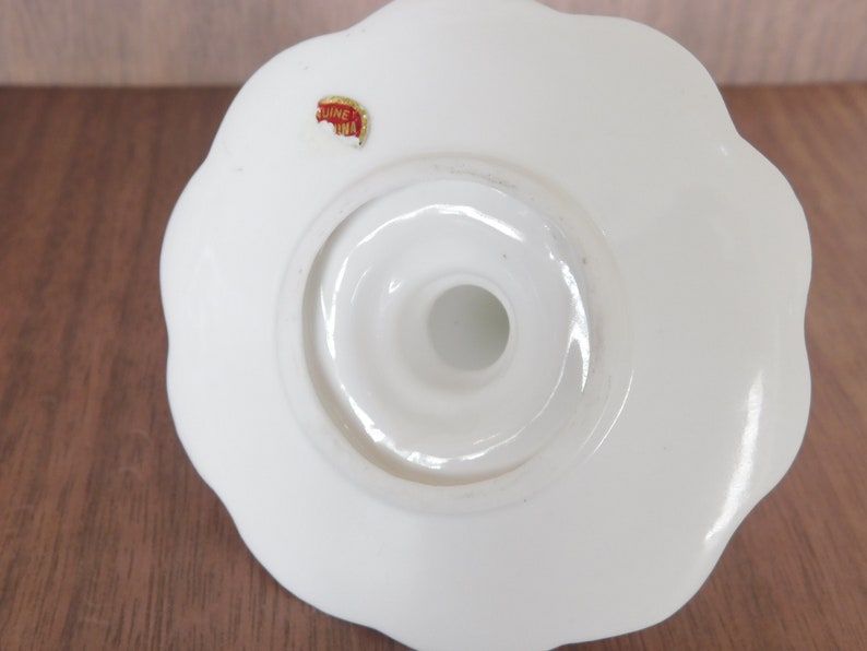 May include: A white ceramic candle holder with a scalloped edge. The base has a circular opening and a small sticker that reads "Genuine China". The candle holder is designed to hold a single candle.