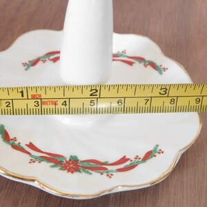 May include: A white porcelain Christmas candle holder with a scalloped edge, gold trim, and festive red and green ribbon and floral design. The holder is approximately 4 inches wide.