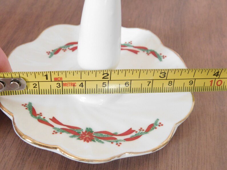 May include: A white porcelain Christmas candle holder with a scalloped edge, gold trim, and festive red and green ribbon and floral design. The holder is approximately 4 inches wide.
