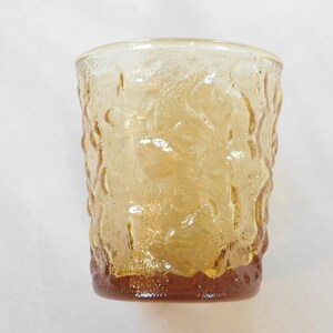May include: A clear amber glass tumbler with a textured, raised design. The glass has a rounded bottom and a slightly flared top.
