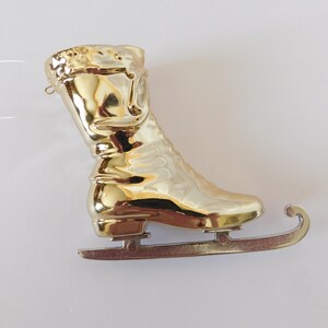 Puede incluir: Adorno de pat&iacute;n de hielo de color dorado con una cuchilla plateada. El pat&iacute;n est&aacute; dise&ntilde;ado para parecerse a un pat&iacute;n de hielo tradicional con una bota y una cuchilla.