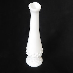 May include: A white milk glass bud vase with a textured, dotted design. The vase has a narrow neck and a wide base.