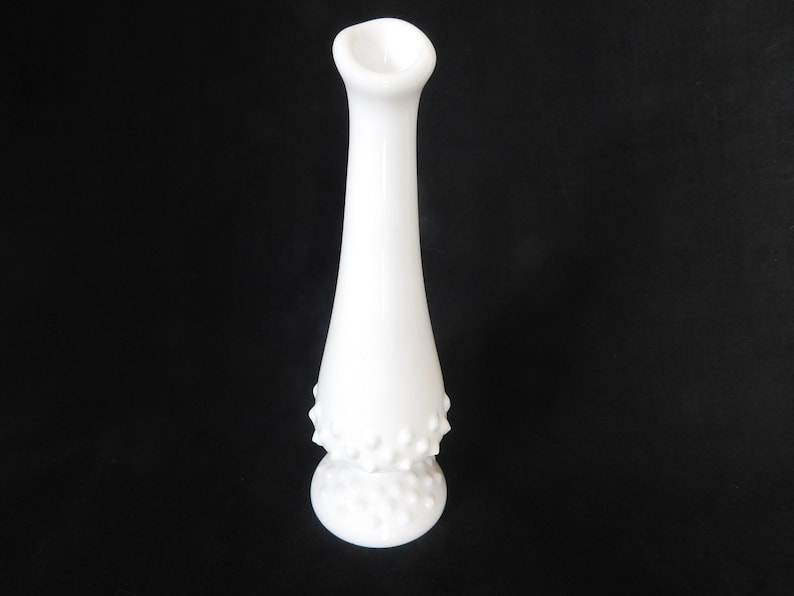 May include: A white milk glass bud vase with a textured, dotted design. The vase has a narrow neck and a wide base.
