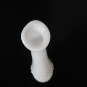 May include: White milk glass bud vase with a beaded design around the bottom. The vase has a flared opening.