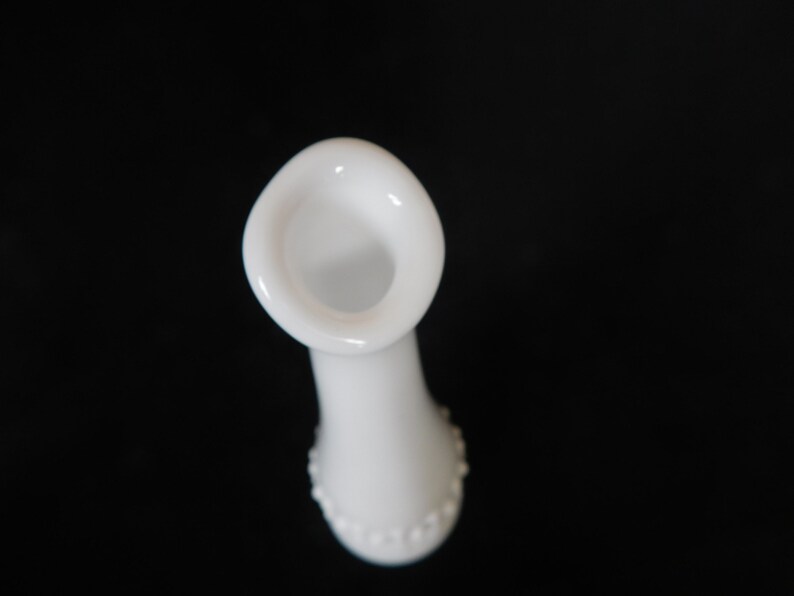 May include: White milk glass bud vase with a beaded design around the bottom. The vase has a flared opening.