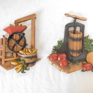 May include: Two vintage wall plaques depicting scenes of rural life. The first plaque features a wooden corn mill with a red wheel, a burlap sack, and a basket of corn. The second plaque shows a black cast iron apple press with a wooden barrel, apples, and a brown jug.