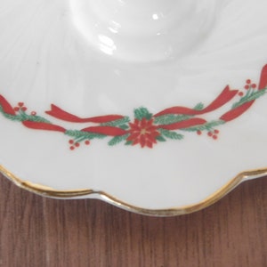 May include: White ceramic dish with a scalloped edge and gold trim. The dish is decorated with a festive design of red ribbons, green pine needles, and red berries, centered around a red poinsettia flower. The dish has a circular indentation.