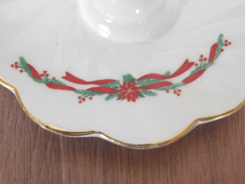 May include: White ceramic dish with a scalloped edge and gold trim. The dish is decorated with a festive design of red ribbons, green pine needles, and red berries, centered around a red poinsettia flower. The dish has a circular indentation.