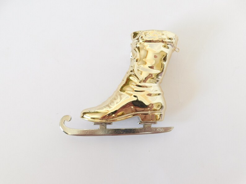 Puede incluir: Adorno de pat&iacute;n de hielo de color dorado con un dise&ntilde;o de bota detallado y una cuchilla de metal plateado.