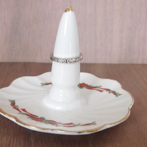May include: White ceramic ring holder with a scalloped base, featuring a gold rim and festive red and green floral accents. A silver ring with clear stones is displayed on the holder. The ring holder is a decorative item.