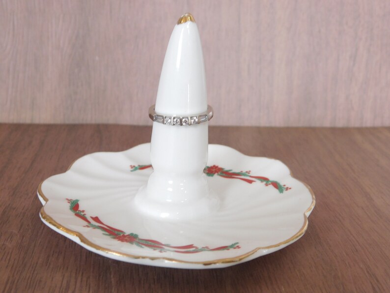 May include: White ceramic ring holder with a scalloped base, featuring a gold rim and festive red and green floral accents. A silver ring with clear stones is displayed on the holder. The ring holder is a decorative item.