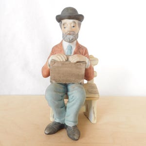 May include: A ceramic figurine of a seated man wearing a hat, jacket, and pants. The man is holding a brown suitcase and is seated on a wooden chair. The figurine is painted in muted colors.