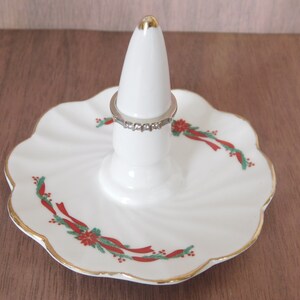 May include: White ceramic ring holder with a gold-trimmed, scalloped edge base. The base features a festive red and green ribbon and floral design. A silver ring with clear stones sits on the holder.