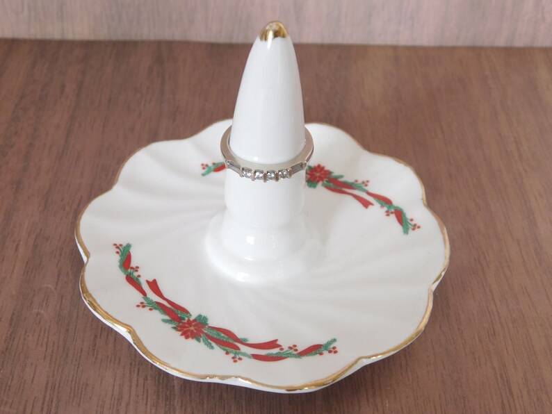 May include: White ceramic ring holder with a gold-trimmed, scalloped edge base. The base features a festive red and green ribbon and floral design. A silver ring with clear stones sits on the holder.