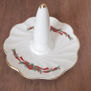 May include: White ceramic ring holder with a gold-trimmed, scalloped edge. The ring holder features a central cone for rings and is decorated with red ribbon and green holly accents, perfect for holiday decor.