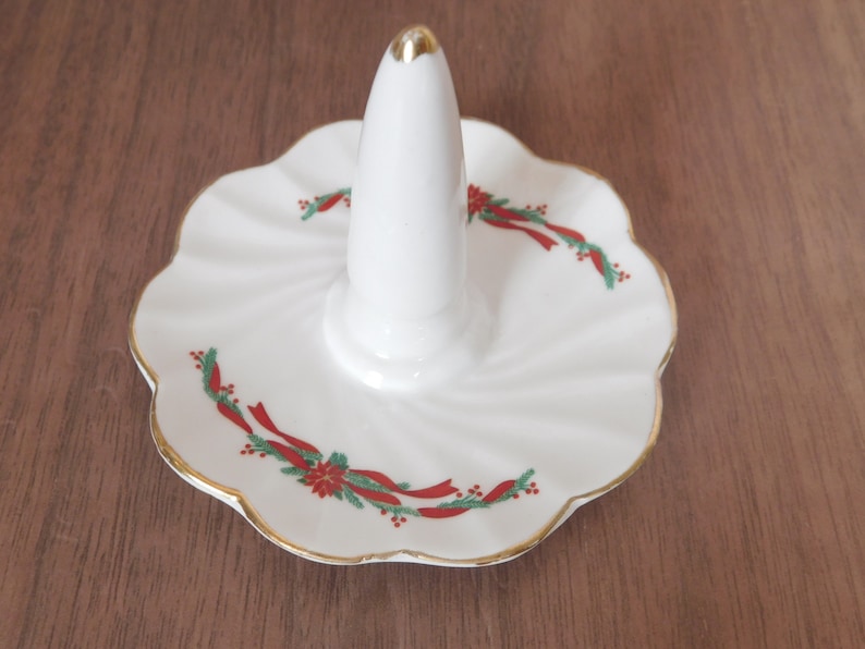 May include: White ceramic ring holder with a gold-trimmed, scalloped edge. The ring holder features a central cone for rings and is decorated with red ribbon and green holly accents, perfect for holiday decor.