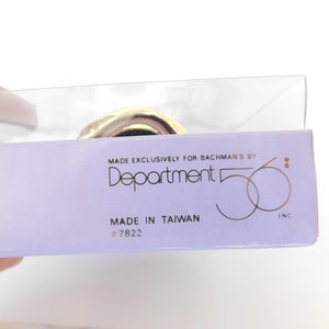Puede incluir: Primer plano de una etiqueta de producto p&uacute;rpura y blanca con el texto "Made Exclusively for Bachman's by Department 56 Inc." y "Made in Taiwan # 7822".