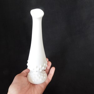 May include: A white milk glass bud vase with a textured, dotted design. The vase has a narrow neck and a wide base.
