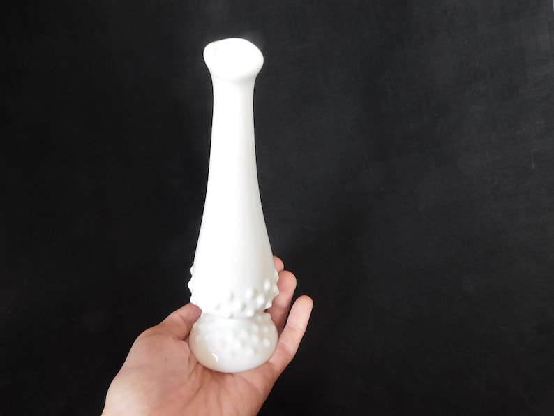May include: A white milk glass bud vase with a textured, dotted design. The vase has a narrow neck and a wide base.