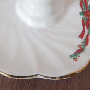 May include: White ceramic candle holder with a scalloped edge and gold trim. The holder is decorated with red ribbons, green pine needles, and red berries, creating a festive holiday design.