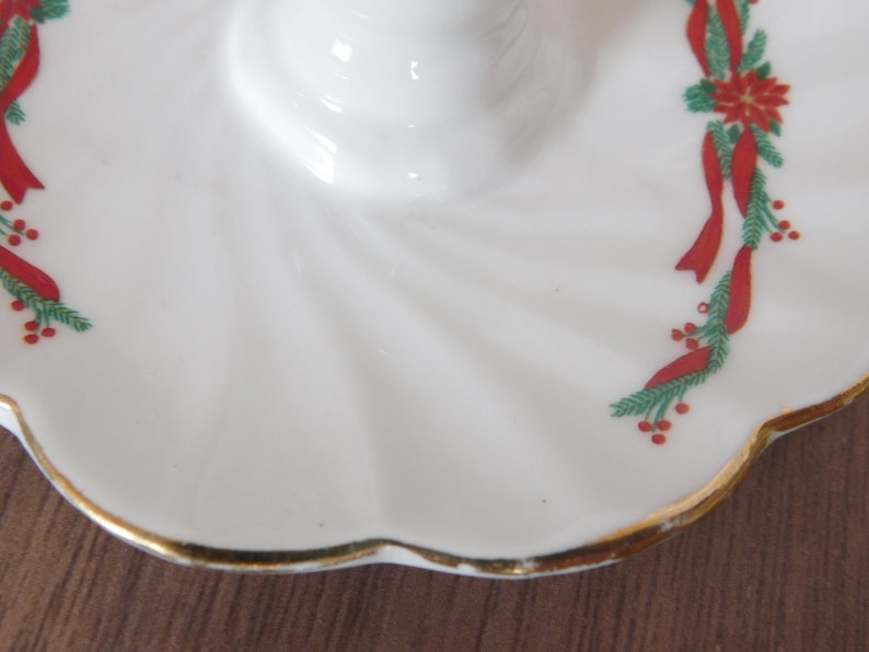 May include: White ceramic candle holder with a scalloped edge and gold trim. The holder is decorated with red ribbons, green pine needles, and red berries, creating a festive holiday design.