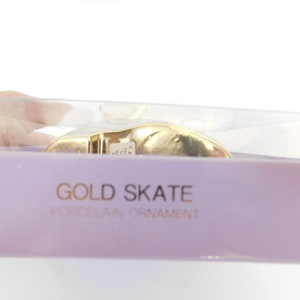 Puede incluir: Un adorno de patinaje de porcelana de color dorado en una caja de pl&aacute;stico transparente con fondo morado. La caja tiene el texto "GOLD SKATE PORCELAIN ORNAMENT" impreso en ella.