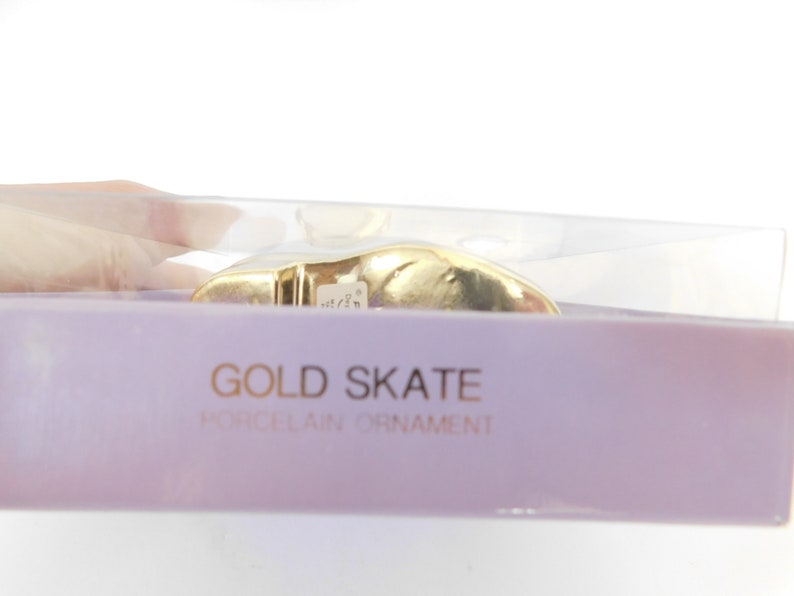 Puede incluir: Un adorno de patinaje de porcelana de color dorado en una caja de pl&aacute;stico transparente con fondo morado. La caja tiene el texto "GOLD SKATE PORCELAIN ORNAMENT" impreso en ella.