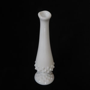 May include: A white milk glass bud vase with a textured, dotted design. The vase has a narrow neck and a wide base.