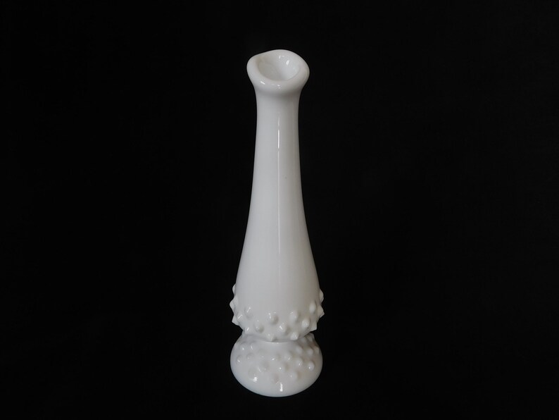 May include: A white milk glass bud vase with a textured, dotted design. The vase has a narrow neck and a wide base.