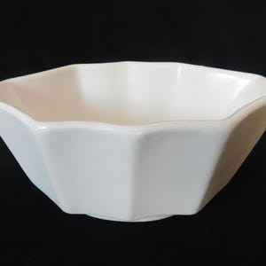 May include: A white, octagonal ceramic bowl. The bowl has a geometric design with eight sides and a slightly flared rim. The bowl is empty and sits against a black background. The bowl is likely for serving food.