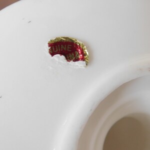 May include: Close-up of a white ceramic object with a glossy finish. A red and gold sticker with the text "GENUINE CHINA" is affixed to the surface. The object has a curved shape with a central opening.