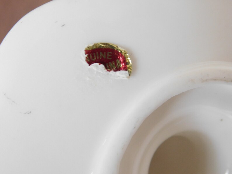 May include: Close-up of a white ceramic object with a glossy finish. A red and gold sticker with the text "GENUINE CHINA" is affixed to the surface. The object has a curved shape with a central opening.
