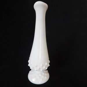 May include: A white milk glass bud vase with a textured, dotted design. The vase has a narrow neck and a wide base.