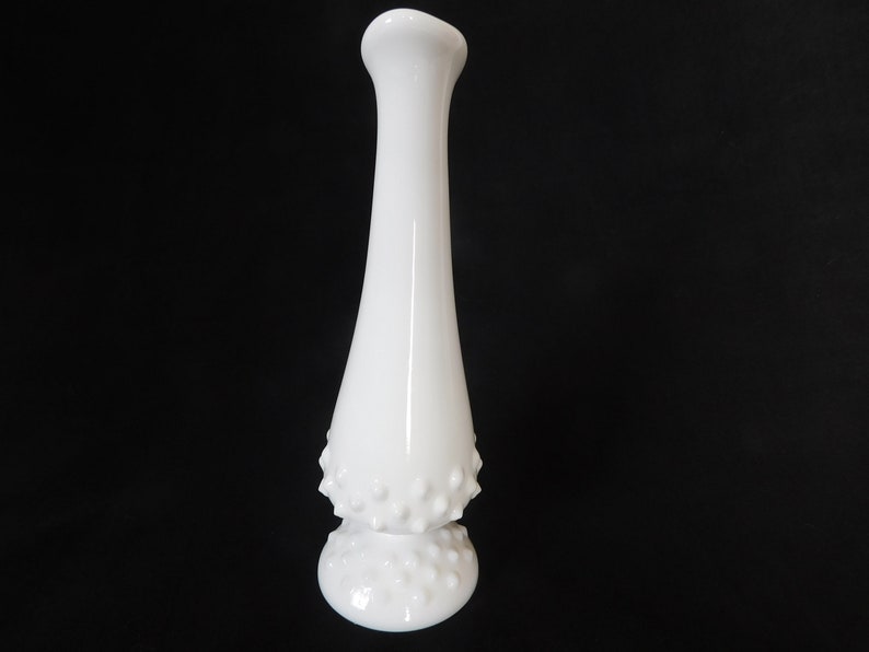 May include: A white milk glass bud vase with a textured, dotted design. The vase has a narrow neck and a wide base.