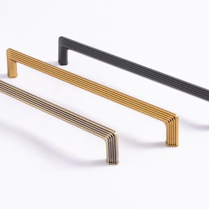 May include: Three cabinet pulls in different finishes: brushed nickel, brass, and black. Each pull has a rectangular shape with a grooved design.