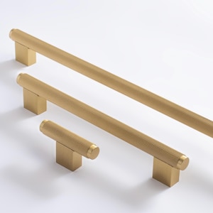 May include: A collection of three brass cabinet pulls in different sizes. The pulls feature a grooved, cylindrical handle design and are mounted on square bases. The hardware is presented against a white backdrop.