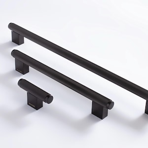 May include: Three black cabinet pulls of varying sizes. Each pull has a cylindrical, textured handle and a rectangular base. The pulls are arranged on a white surface, showcasing their design and potential for use in home decor.