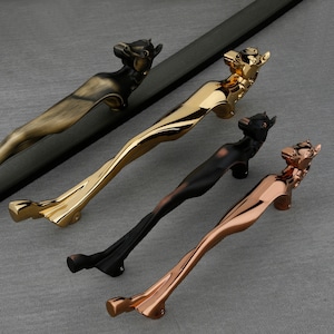 May include: Four decorative door handles in various metallic finishes: bronze, gold, black, and copper. Each handle features a stylized animal head at the top and a curved, ergonomic design. The handles are arranged on a gray surface.