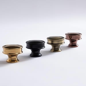 May include: Four decorative cabinet knobs in gold, black, brass, and copper. Each knob has a rounded top and a cylindrical base. The knobs are arranged in a row against a white background, showcasing their different metallic finishes.