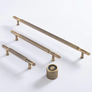 May include: A set of four antique brass cabinet hardware items. The set includes three bar pulls of varying lengths and a round knob. Each piece has a ribbed design and T-shaped ends. The items are displayed on a white background.
