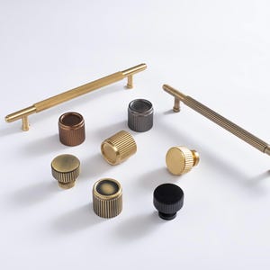 May include: A collection of brass and metal cabinet hardware, including handles and knobs. The handles have a ribbed design, and the knobs come in various colors, including gold, bronze, and black. The hardware is arranged on a white surface.