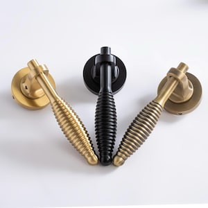 以下が含まれることがあります： 真鍮と黒の装飾的なドアハンドル3つ。ハンドルはリブのデザインで、丸いバックプレートに取り付けられています。真鍮のハンドルは暖かく金色の色合いで、中央のハンドルは黒です。ハンドルは白い背景に配置されています。