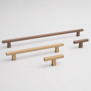 Könnte beinhalten: Eine Sammlung von Schrankgriffen und -knöpfen in Bronze- und Goldtönen. Die Griffe sind rechteckig mit gerippter Textur, und die Knöpfe haben eine T-Form. Die Hardware wird vor einem weißen Hintergrund präsentiert und zeigt die verschiedenen Größen und Ausführungen.